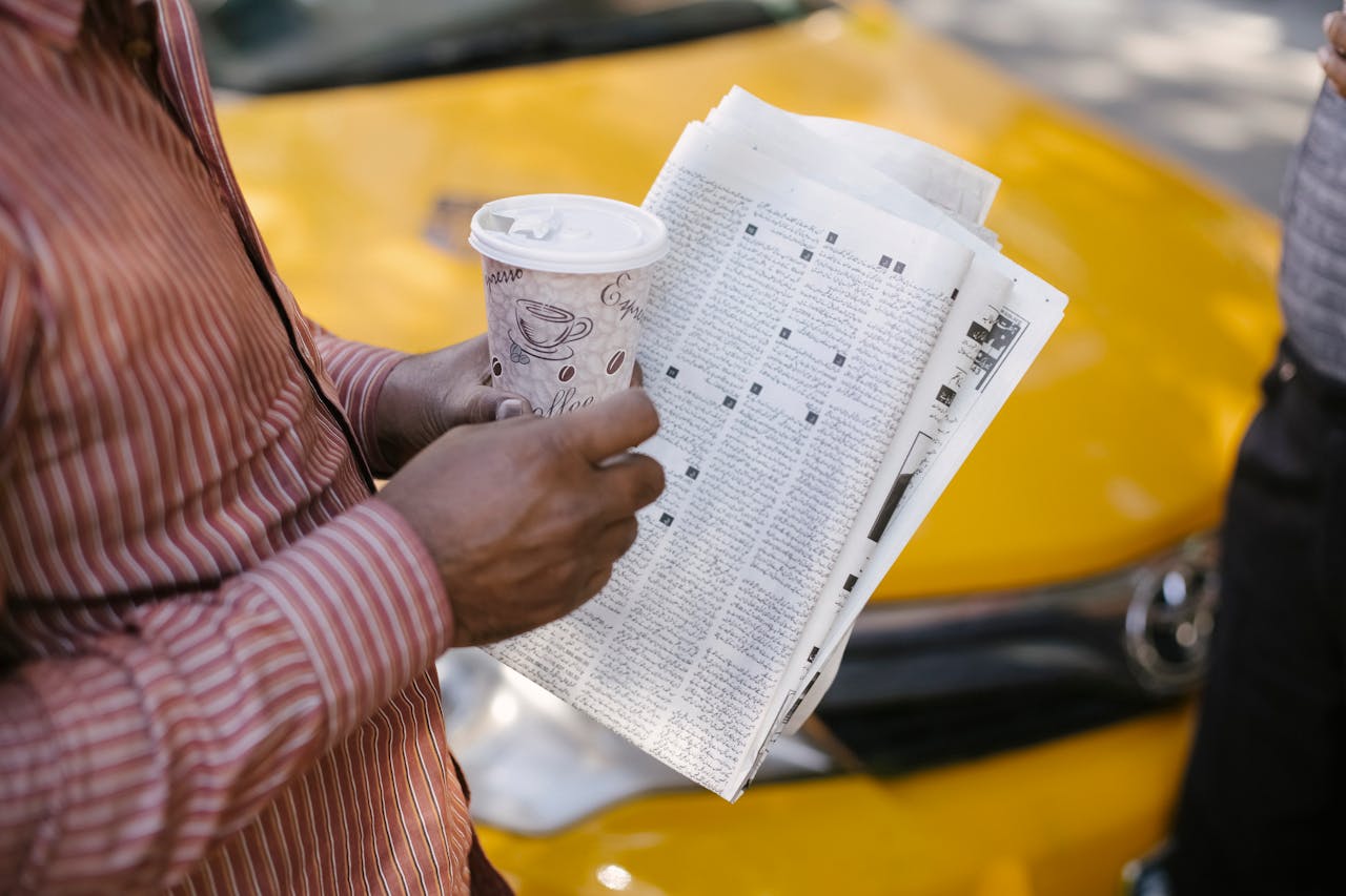 From above side view of crop anonymous ethnic male cab driver with coffee to go and newspaper talking to coworker on street